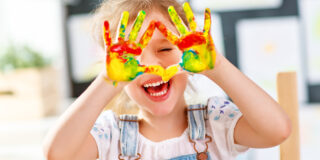 Ein Kind lacht und zeigt mit Händen ein Herz, die Hände sind mit Fingerfarbe bunt bemalt.