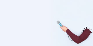 Eine Hand hält ein Mikro vor blanker Wand.