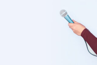 Eine Hand hält ein Mikro vor blanker Wand.