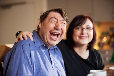 Zwei Menschen - ein Mann und eine Frau - sitzen vertraut und lachend am Tisch. Die Frau legt dem Mann eine Hand auf die Schulter.