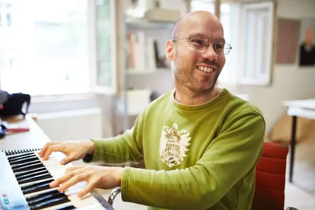 Ein junger Mann mit Brille spielt Klavier und lächelt.