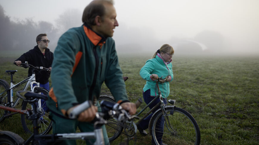 Drei Menschen - behindert und  nicht behindert -  schieben ihre Fahrräder in morgendlichem Dunst an einer Wiese.