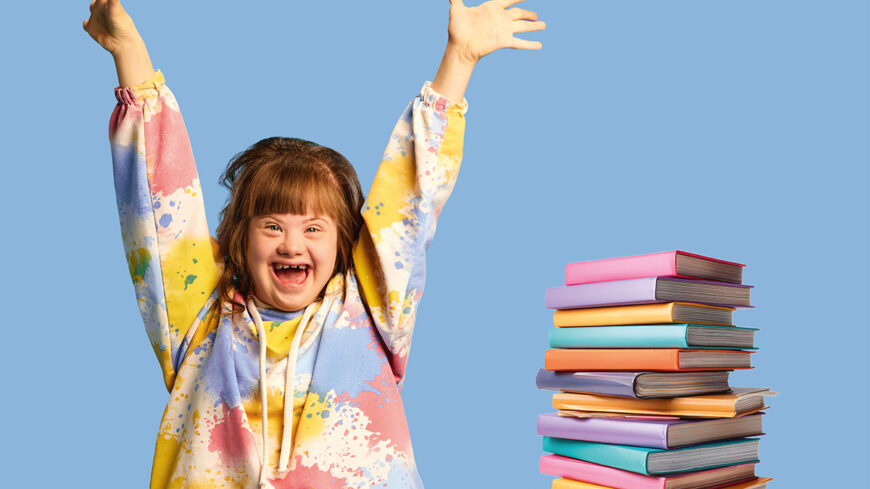 Ein Mädchen mit Down-Syndrom trägt einen bunten Jogginganzug und freut sich, neben ihr steht ein bunter Stapel Bücher.